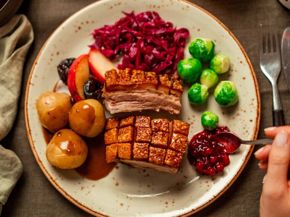 En tallerken med kokte poteter, rødkål, skiver av svinekjøtt, sprø svineflesk og tyttebærsyltetøy med en skje som holder tyttebær.