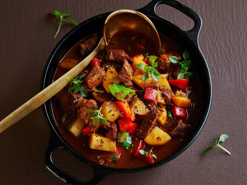 Gulasj i en svart gryte med kjøtt, poteter, gulrøtter og paprika, pyntet med urter.