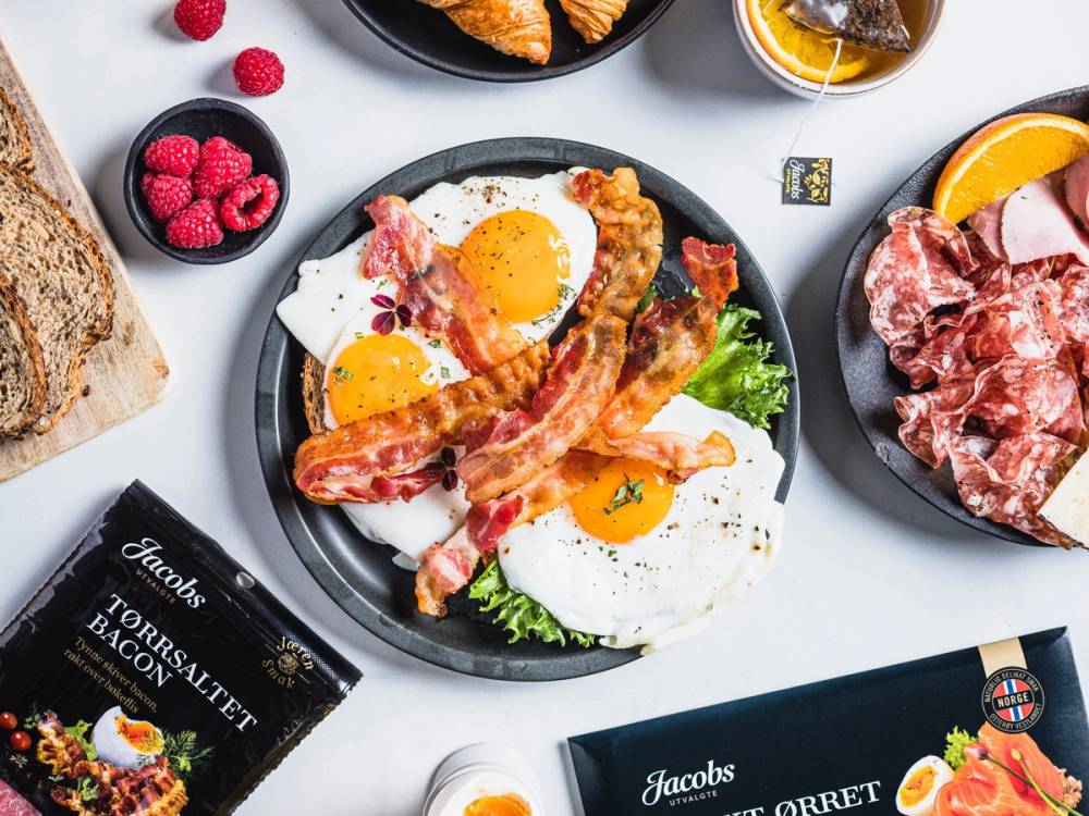 Frokost med stekte egg, bacon, brød, skinke og frukt på et bord.