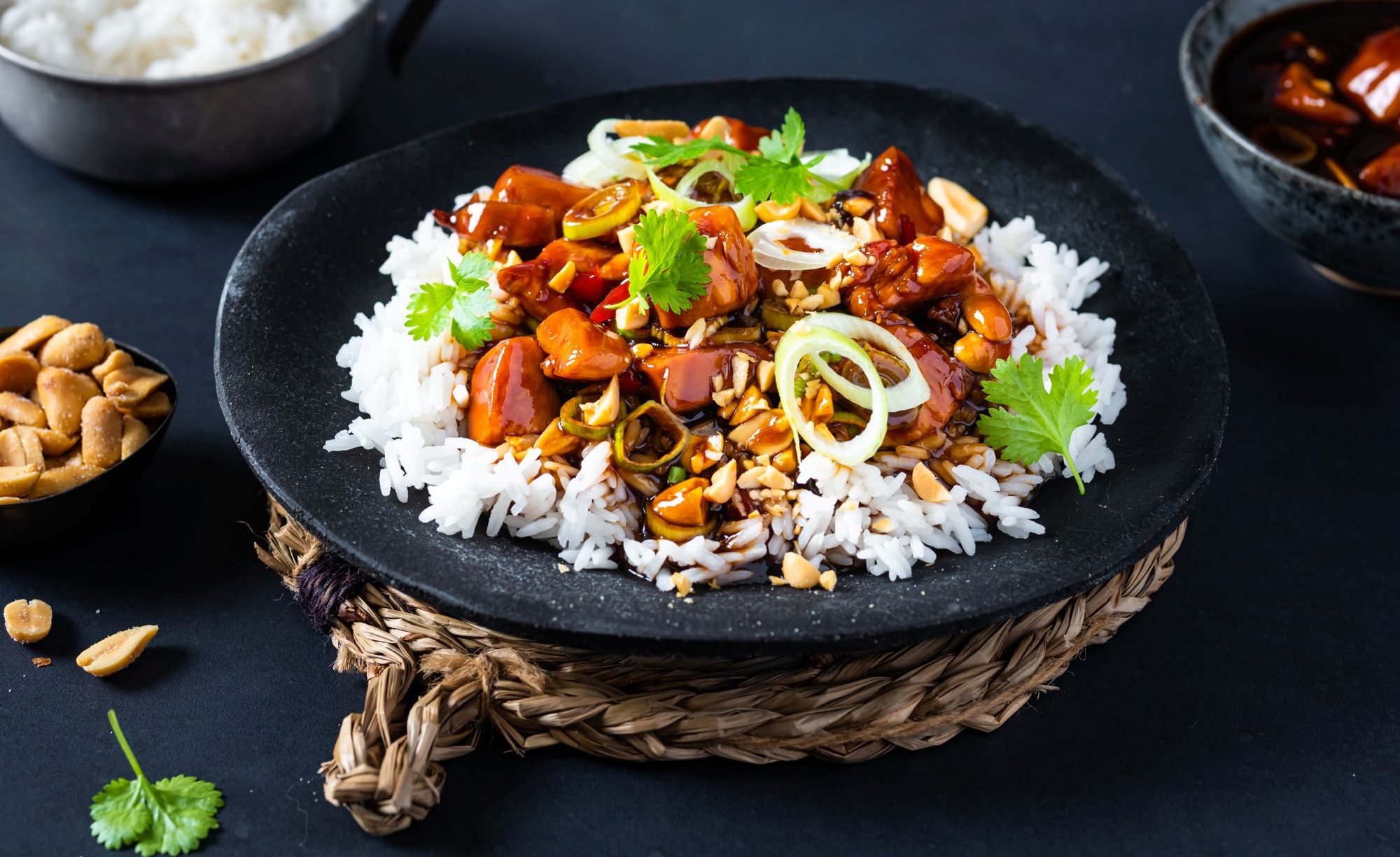 Kylling Gong Bao servert med ris og garnityr av nøtter og grønnsaker på en svart tallerken