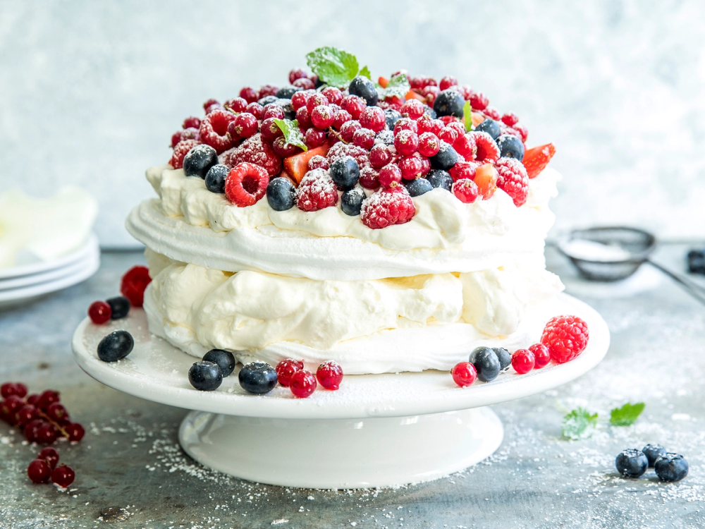 En sommerpavlova dekket med ulike bær på en hvit kakefat, pyntet med friske bær og mynteblader