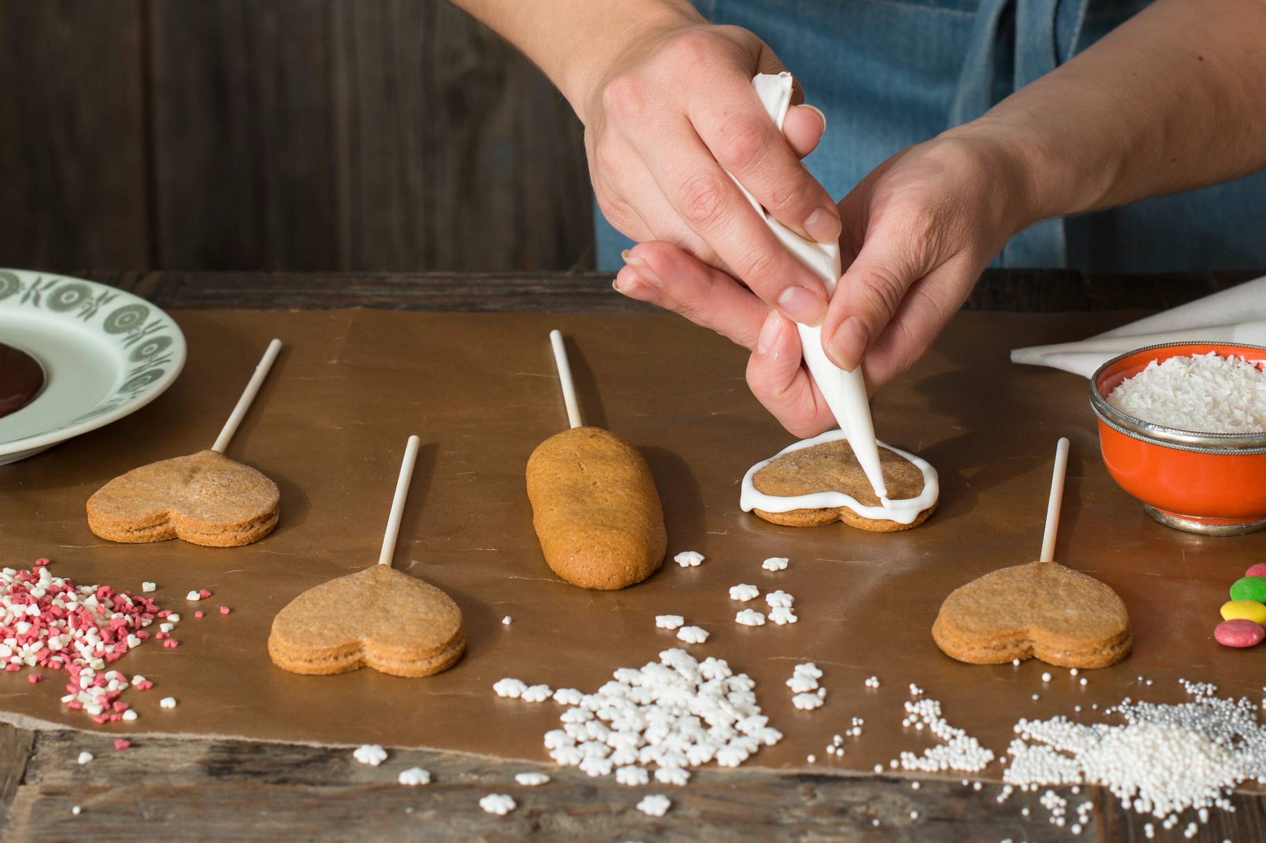 Person som pynter pepperkaker med krem på et trebord med ulike pepperkakeformer og pynt rundt.