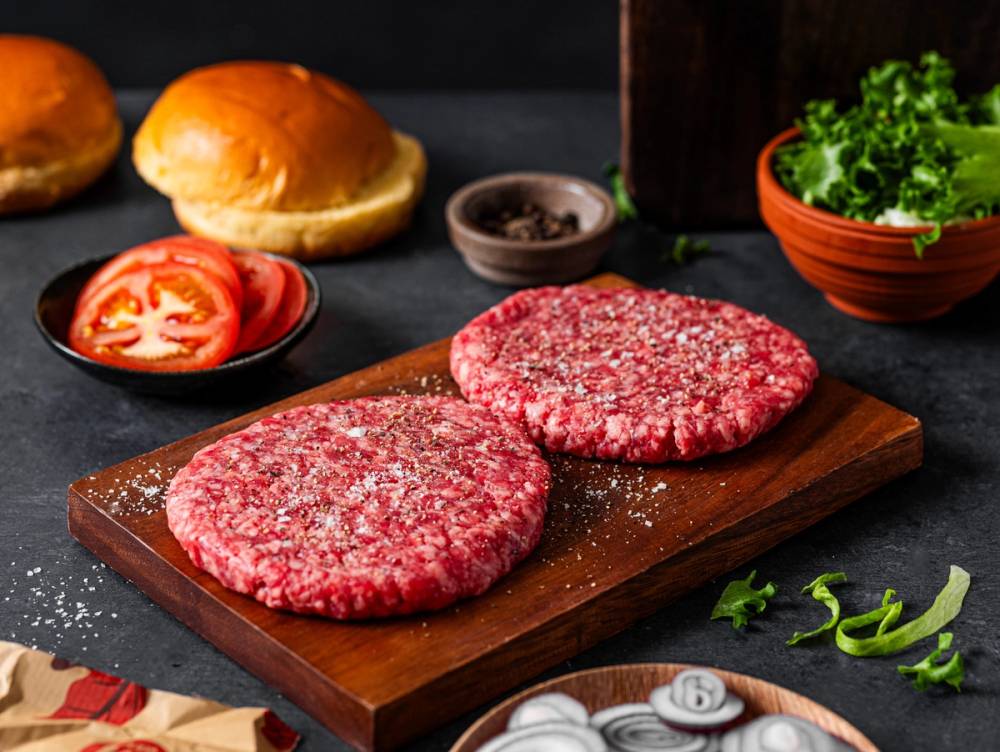 To rå kjøttkaker på en skjærebrett med salat og tomatskiver i bakgrunnen.