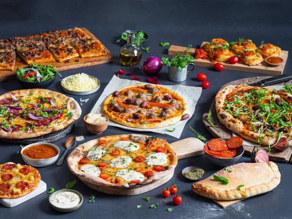 Et utvalg av pizzaer med ulike toppings på et bord, omgitt av små skåler med saus og grønnsaker, klar for helgekos.