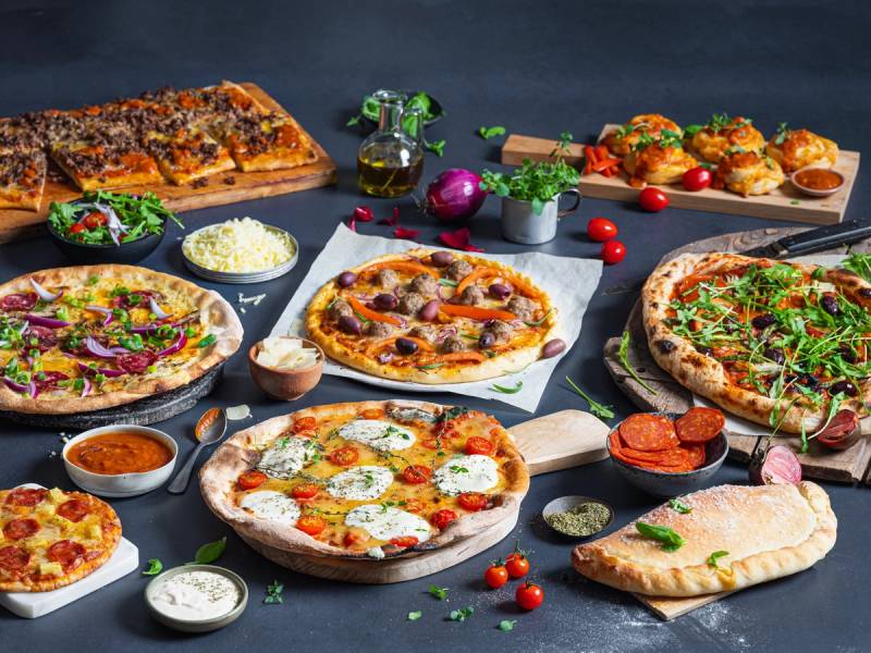 Et utvalg av pizzaer med ulike toppings på et bord, omgitt av små skåler med saus og grønnsaker, klar for helgekos.