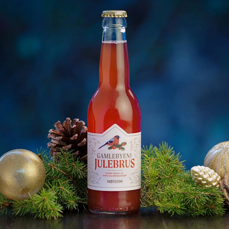 En flaske julebrus plassert på en juledekorasjon med granbar og julepynt, mot en blå bakgrunn.