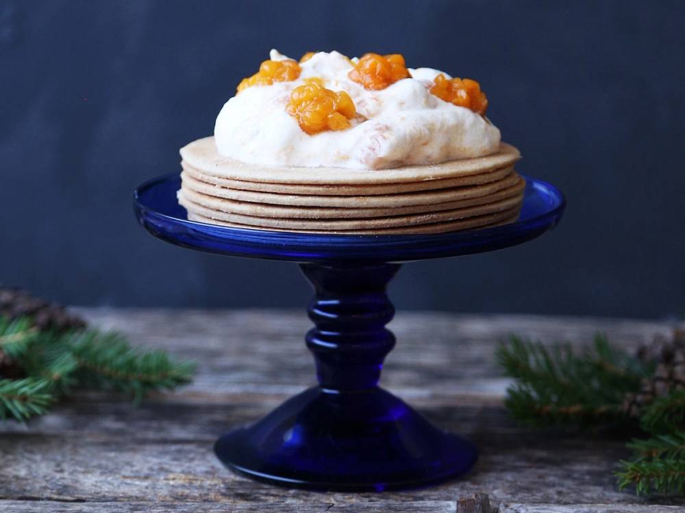 En tallerken med flere tynne pannekaker toppet med krem og kandiserte appelsinbiter, plassert på en blå tallerken på et bord med granbar rundt.