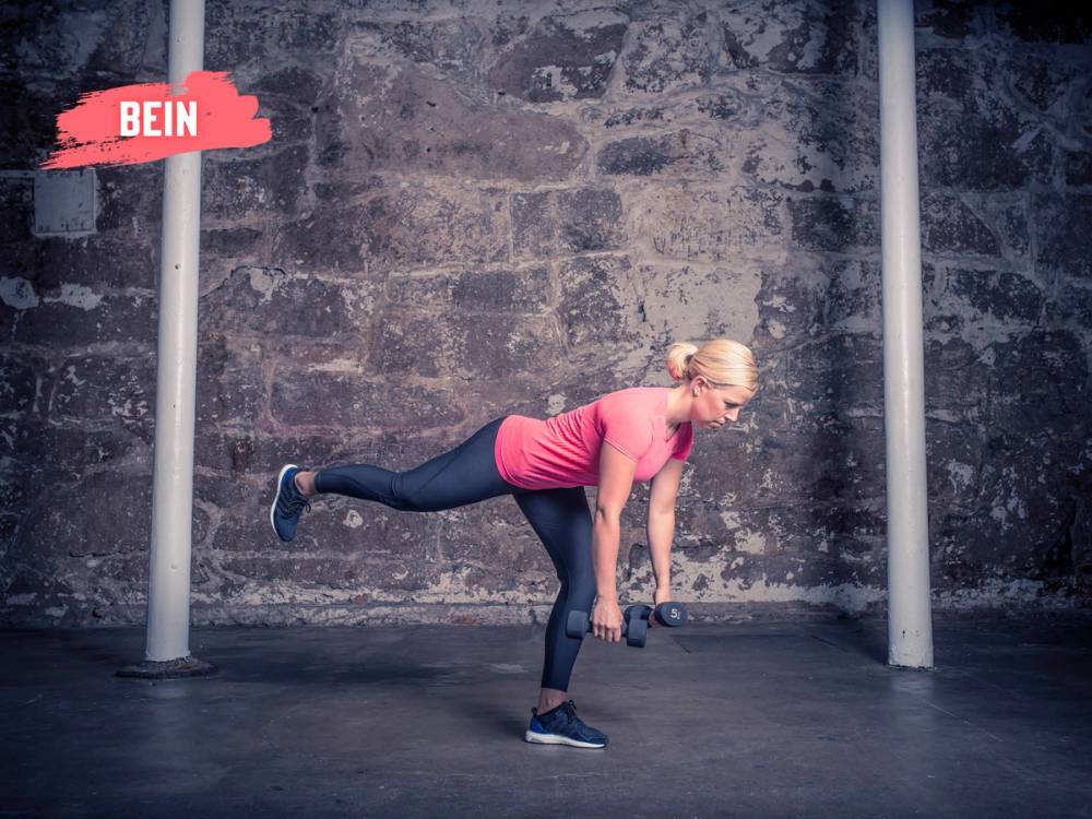 En kvinne utfører en ettbeins strakmark i et treningsrom med en murvegg i bakgrunnen.