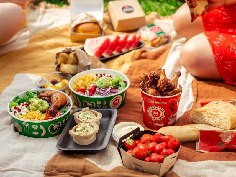 Et utvalg av mat og snacks på et teppe, inkludert salat, frukt, kjøttboller, tomater og pommes frites, med personer som sitter rundt.