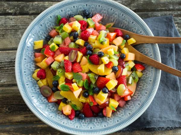 En stor blå bolle fylt med en fargerik fruktsalat med biter av mango, jordbær, blåbær, druer og kiwi, med en tresleiv oppi.