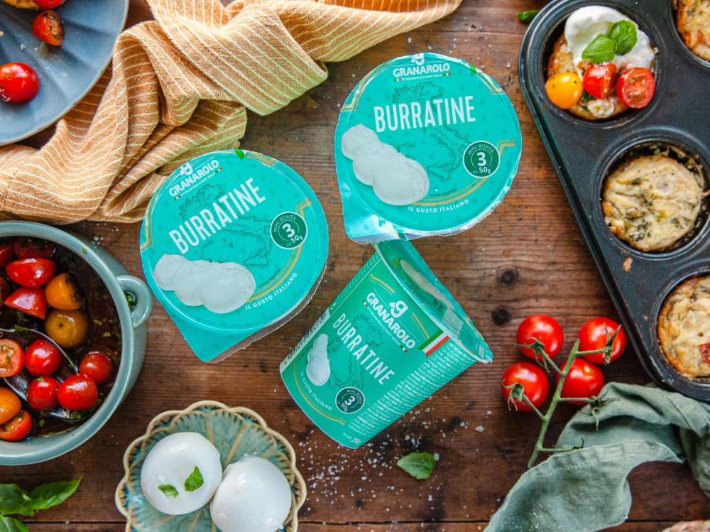 To små bokser med burbætte, omgitt av cherrytomater, et egg, og en skje med mat, på et trebord med en klut og en stekepanne med mat i.