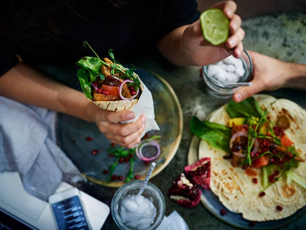 Person som holder en wrap med grønnsaker og kjøtt, omgitt av flere andre wraps og en kake på et bord.