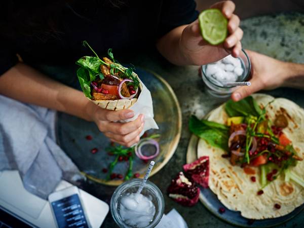 Person som holder en wrap med grønnsaker og kjøtt, omgitt av flere andre wraps og en kake på et bord.