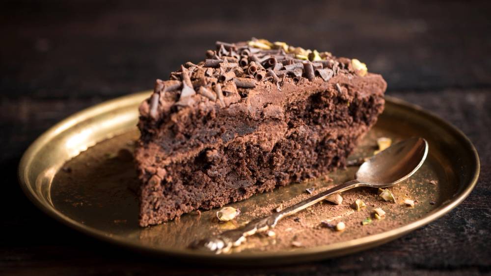 En skive sjokoladekake med sjokoladekrøller på toppen på en gullfarget tallerken med en skje ved siden av