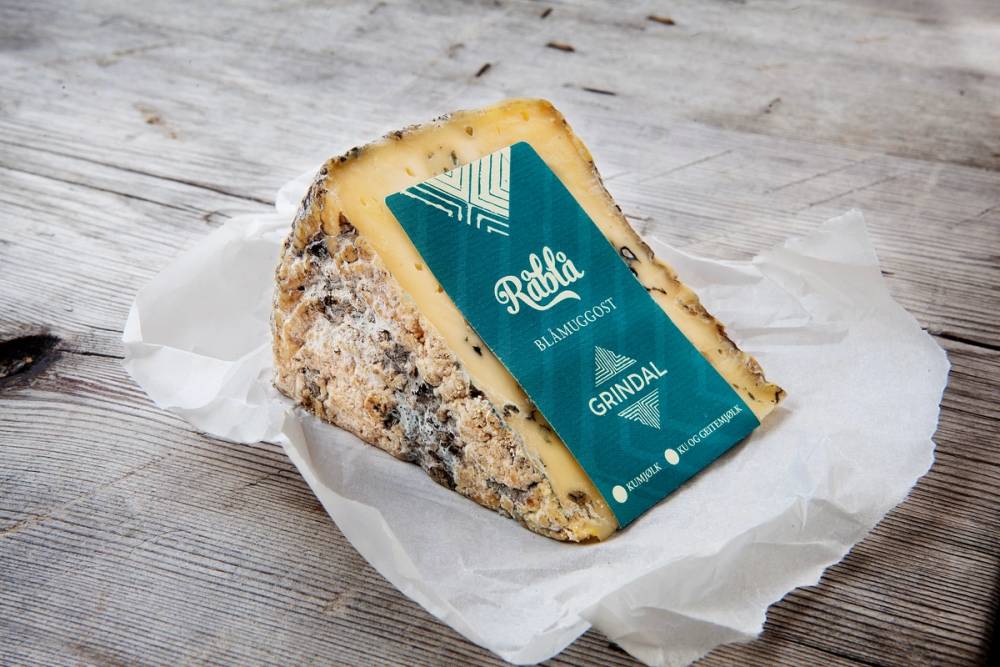 Et stykke råblå ost med blåmuggskorpe ligger på hvitt papir på et lyst trebord, med en etikett som sier Råblå og Grøndal.
