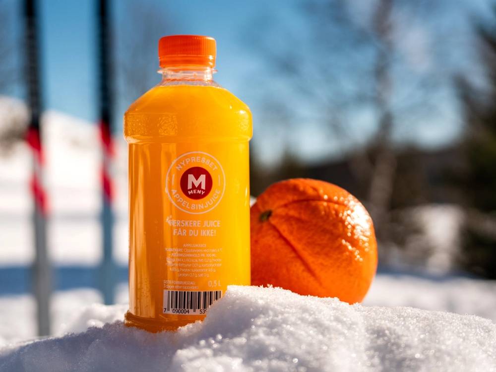 En flaske med oransje drikke og en liten oransje ball ligger på snø utendørs med blå himmel og trær i bakgrunnen.