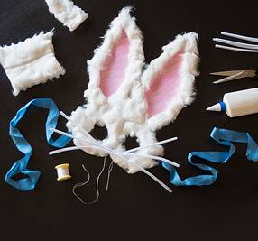 En påskehare-maske laget av hvit pels med rosa innsiden av ørene, omgitt av sytilbehør og verktøy for å lage masker.