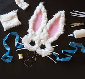 En påskehare-maske laget av hvit pels med rosa innsiden av ørene, omgitt av sytilbehør og verktøy for å lage masker.