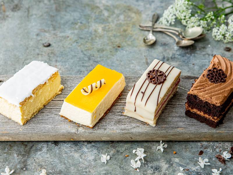 Fire ulike kaker på et trebrett med blomster og bestikk i bakgrunnen.