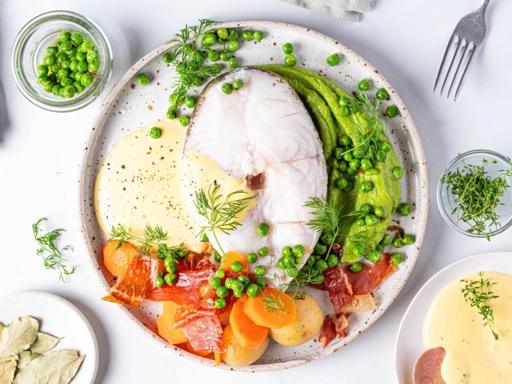 Skrei med spekeskinke, grønne erter, og hollandaise-saus på en hvit tallerken med urter og grønnsaker rundt
