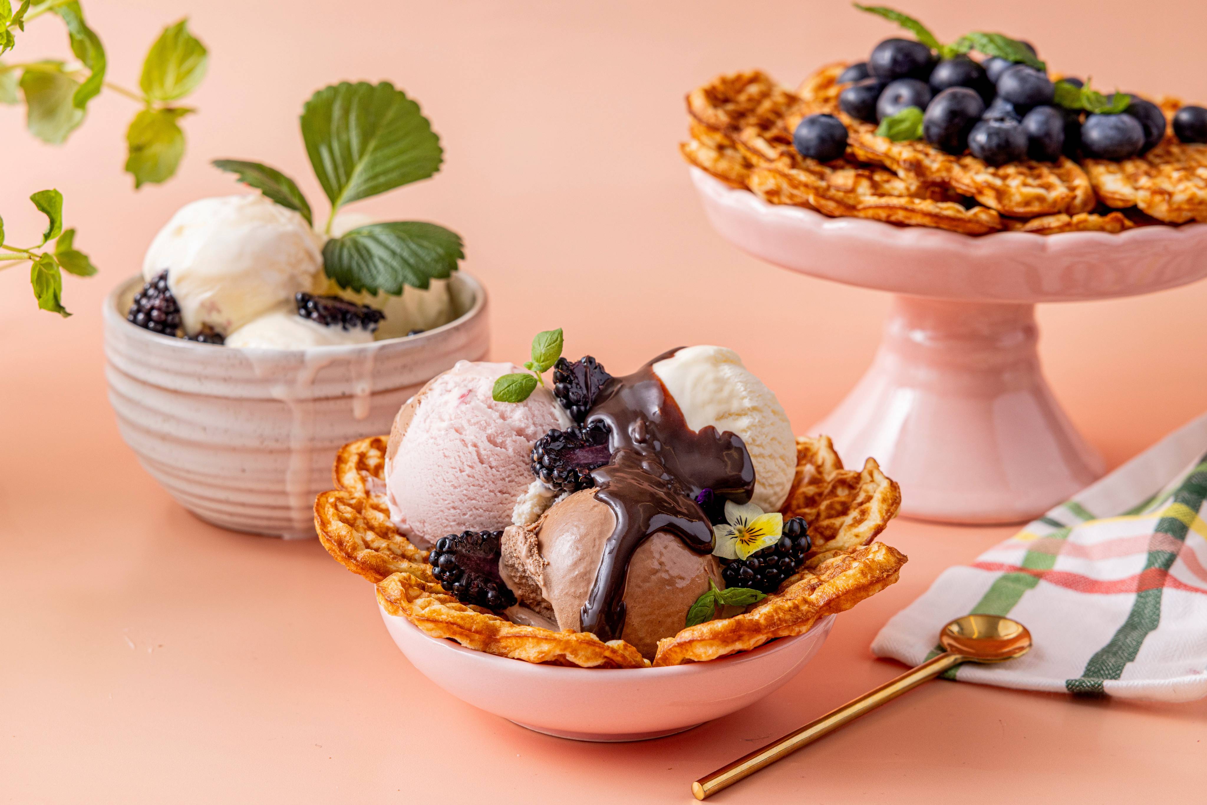 Vafler med iskrem, blåbær og sjokoladesaus, servert på små fat, med en skje og et strikket serviett ved siden av.