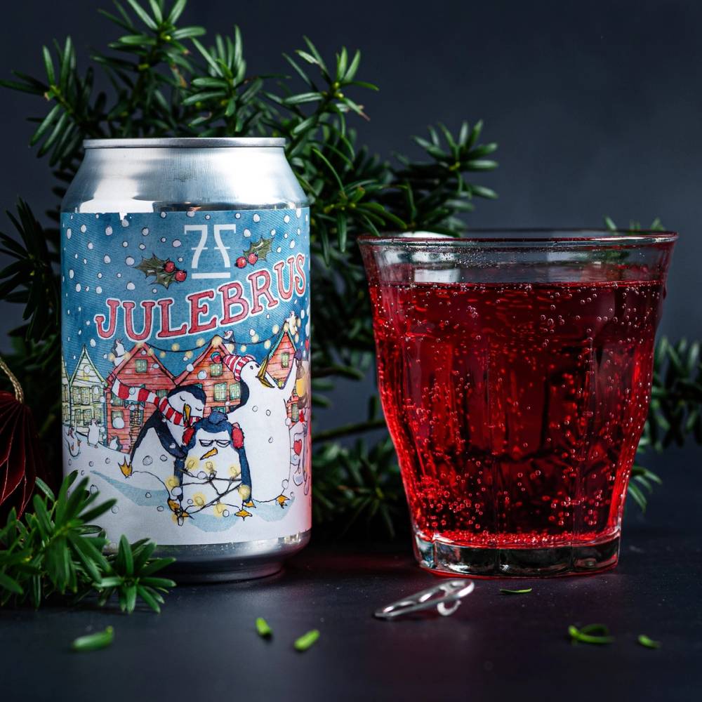 En julebrusboks med julemotiver og en glass med rød julebrus ved siden av grønne grener, mot en mørk bakgrunn.