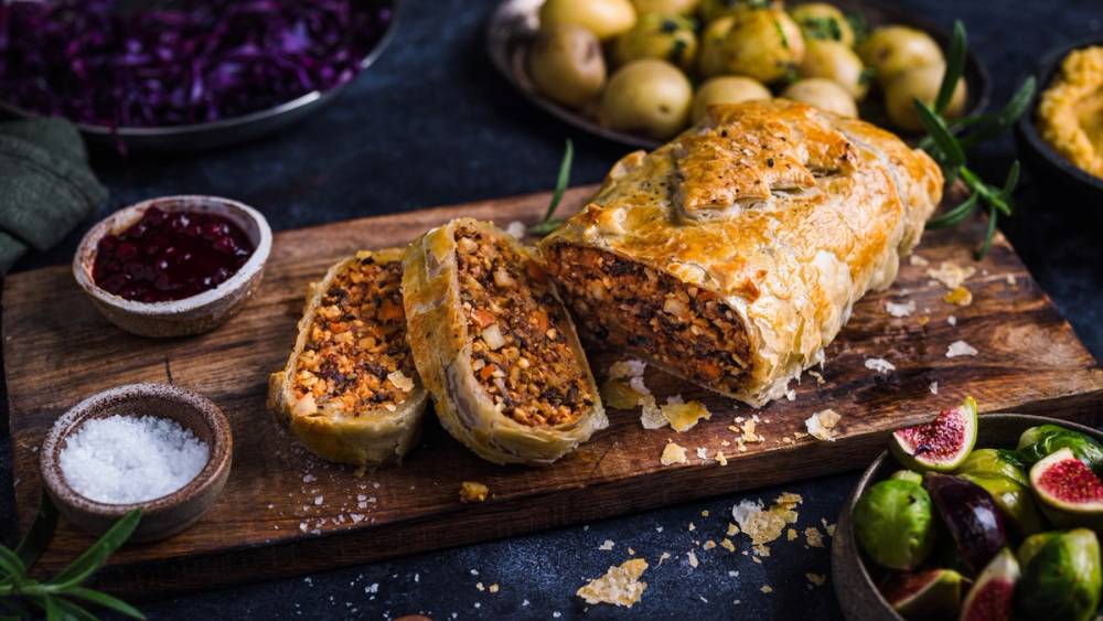 Nøttestek med fylling skåret opp på et skjærebrett, omgitt av små skåler med syltetøy og salt, og plommer i bakgrunnen.