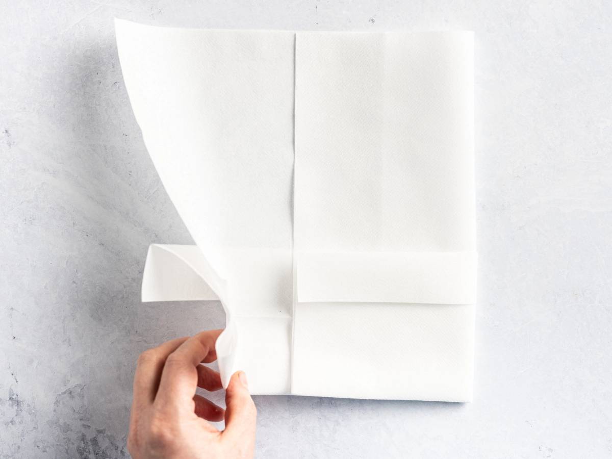 En person holder en brettet hvit serviett med en lomme, klar til bruk på et bord.