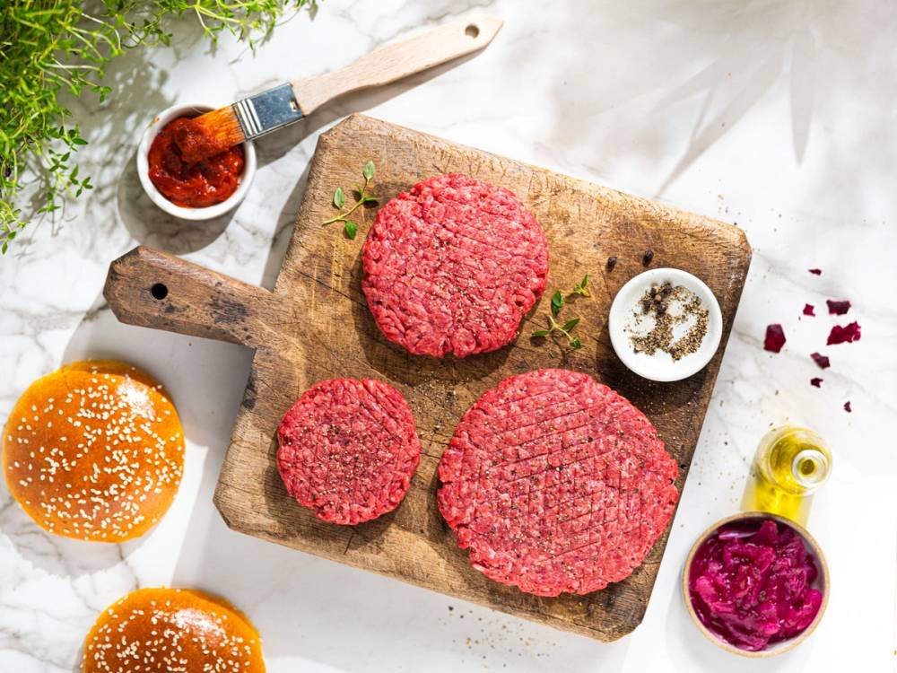 Tre rå Angus-burgere på et skjærebrett med krydder og sauser rundt, klare for grilling eller steking.