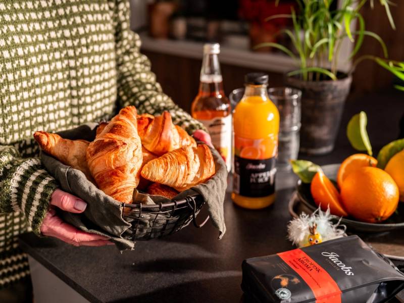 En person holder en svart kurv med ferske croissanter foran en bord med ulike drikker og frukt, inkludert appelsiner og en plante i bakgrunnen.