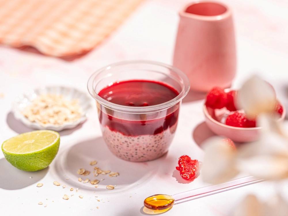 En dessert i et glass med havregryn, chiafrø og rød bærsaus, omgitt av lime, kokosflak og frukt, på et rosa bord.