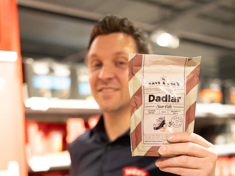 Person holder en pose med tekst og bilder på i en butikk, med butikkhyller i bakgrunnen.