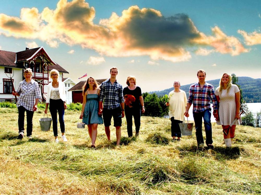 En gruppe mennesker går på en gresslette med hus i bakgrunnen under en solrik himmel med noen skyer.