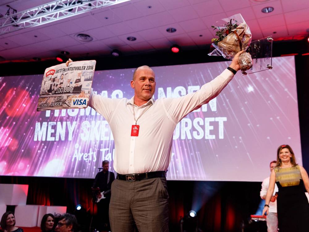 En person med oppkuttet ansikt holder en blomsterbukett og et gavekort på scenen under en prisutdeling, med en annen person i bakgrunnen.