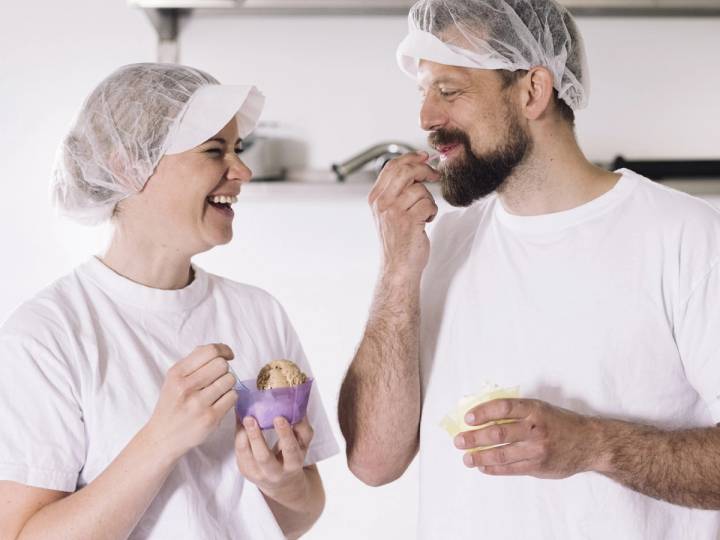 To personer i hvite klær og hårnett spiser iskrem i et kjøkkenmiljø, med fokus på iskremen de holder.