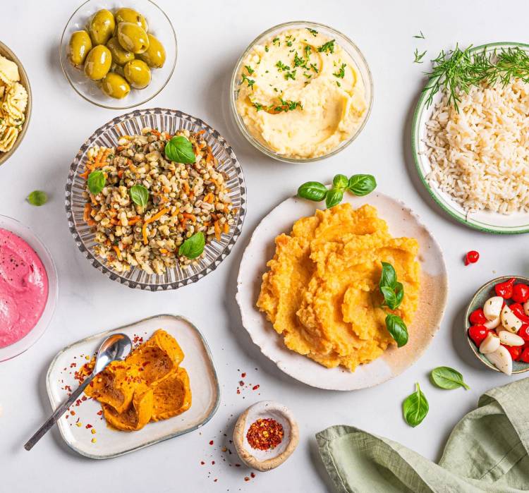 Ulike retter og tilbehør på et bord, inkludert potetmos, ris, grønne oliven, og salat med cherrytomater, med en grønn serviett i hjørnet.