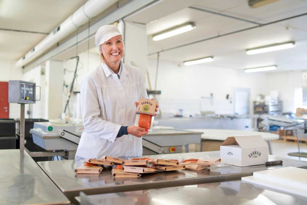 Person i hvit labbekk med ansiktet dekket til, som pakker kjøttpålegg i en industriell produksjonslinje, med flere pakker og utstyr rundt seg.