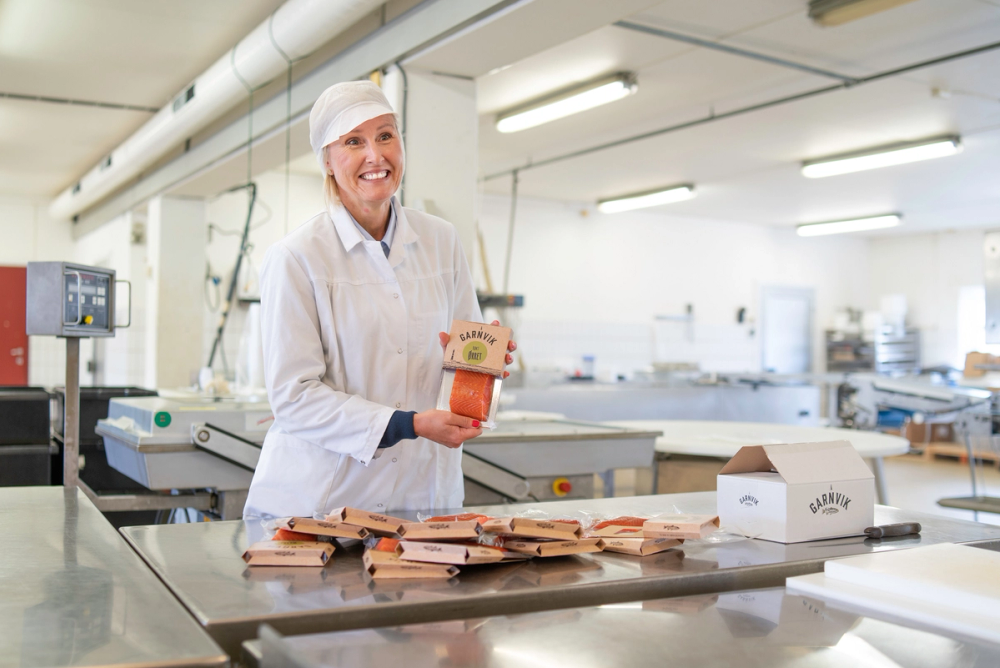 Person i hvit labbekk med ansiktet dekket til, som pakker kjøttpålegg i en industriell produksjonslinje, med flere pakker og utstyr rundt seg.