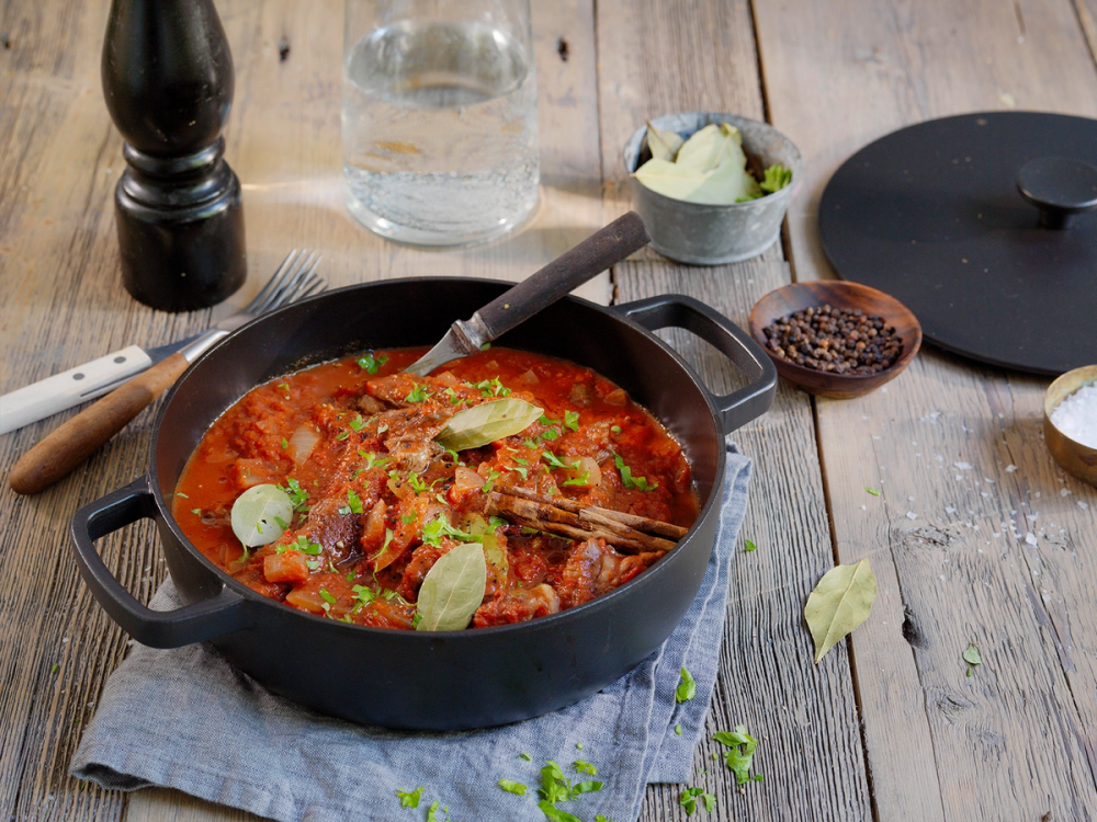 En gryte med stifado, en gresk gryterett med kjøtt og grønnsaker i en tomatsaus, plassert på et trebord med krydder og tilbehør rundt.