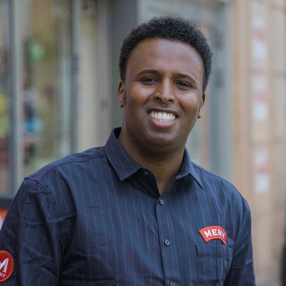 En mann i blå skjorte med logo står utendørs med bygninger i bakgrunnen.