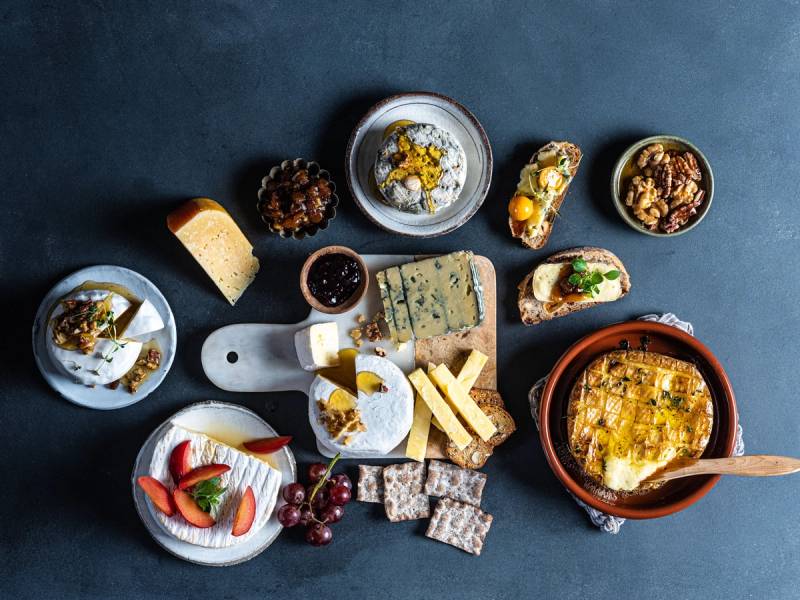 Et ostefat med ulike typer ost, kjeks, frukt og tilbehør på et mørkt bord.