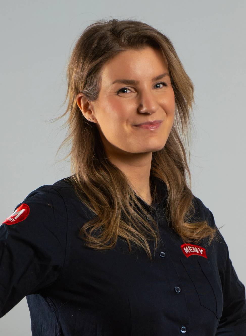 En kvinne med langt, lyst hår iført en svart arbeidsuniform med navnemerke og en logo på ermet, står mot en lys bakgrunn.