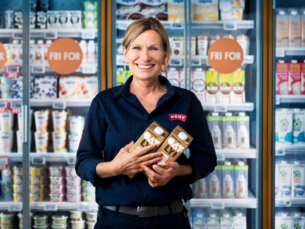 En person i butikkuniform holder to melkekartonger foran kjøleskap med meieriprodukter, med en logo som sier 'Meny' på uniformen.