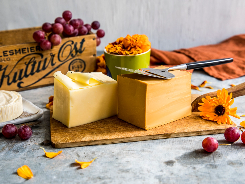 Brunost og gulost på et skjærebrett med blomster og druer rundt, klar til frokost.