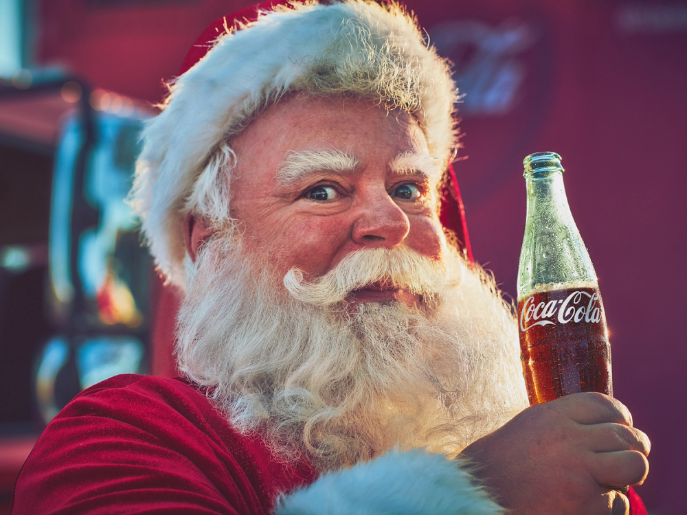 Person kledd i juleantrekk holder en Coca-Cola-flaske under en julepyntet bakgrunn.