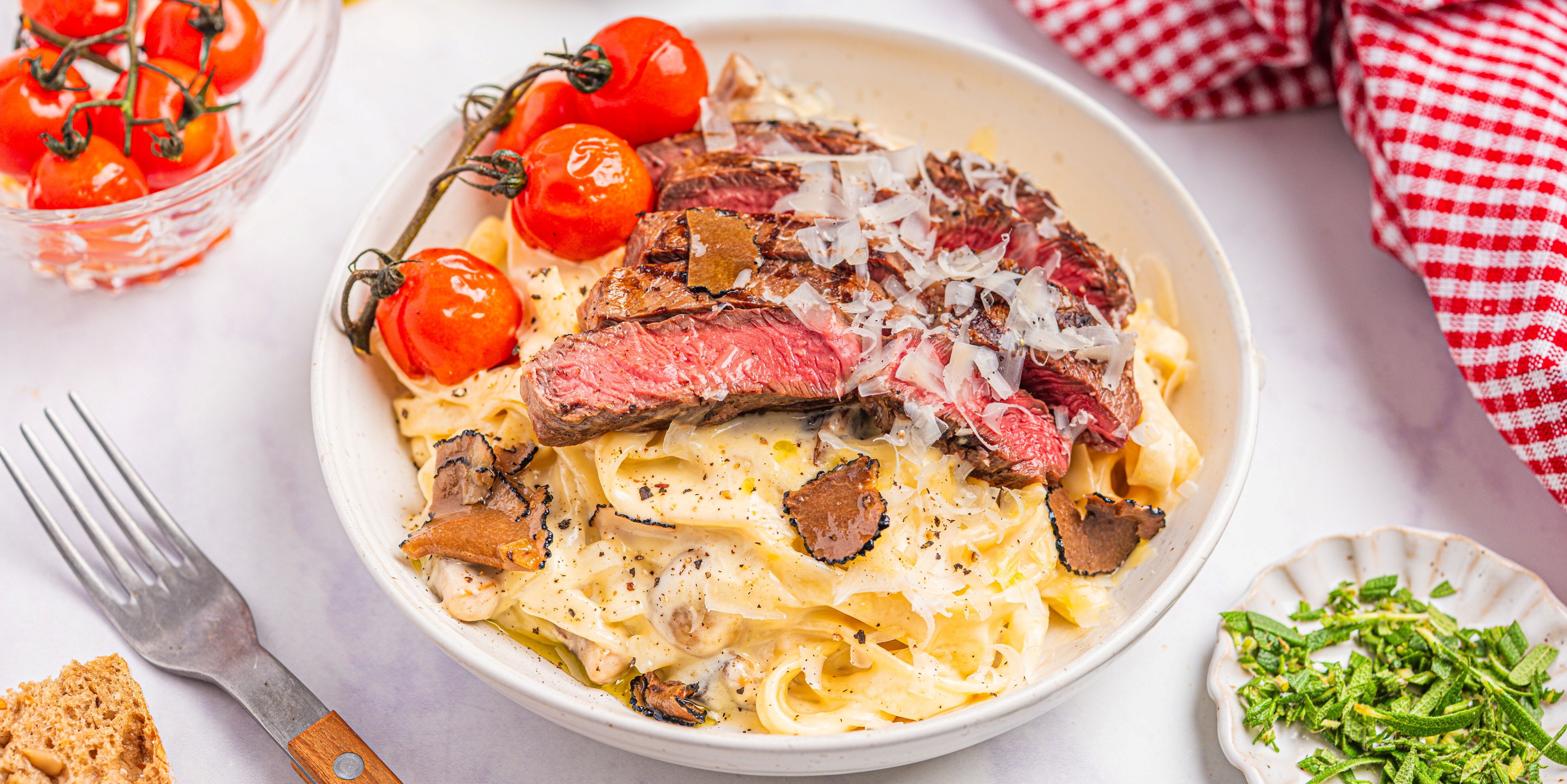 Trøffelpasta med biff og cherrytomater på en hvit tallerken, omgitt av en liten skål med grønne urter og et glass med vann, på et hvitt bord.