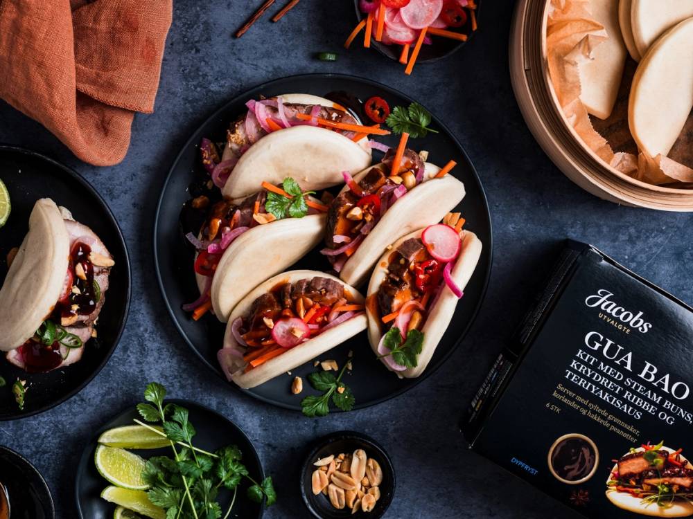 Gua Bao med svinekjøtt, grønnsaker og saus på en svart tallerken, omgitt av andre ingredienser og en pakke med Jacobs Gua Bao, på et mørkt bord.
