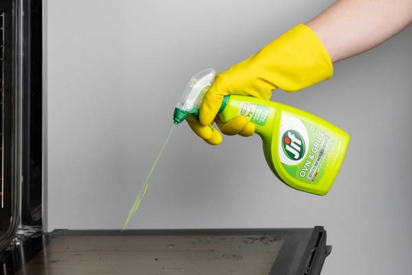 En person med gule hansker sprayer rengjøringsmiddel på en ovn eller grill, som står på en kjøkkenbenk.