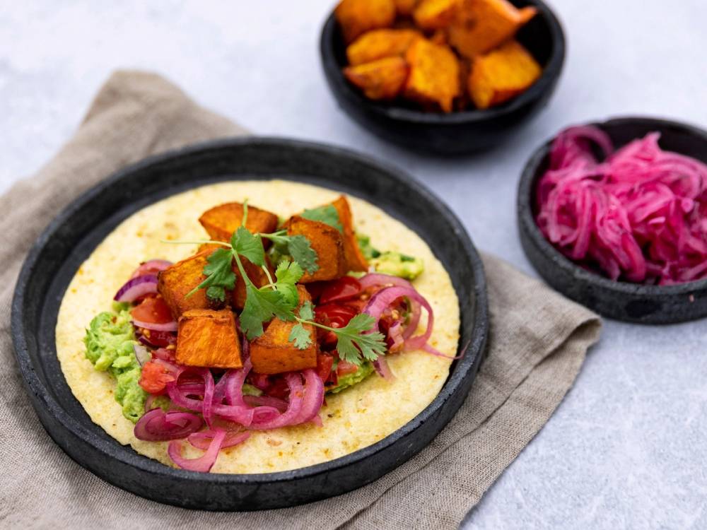 Bakt hokkaidogresskar på taco med guacamole, rødløk og koriander på en tallerken, med to små boller med tilbehør i bakgrunnen.