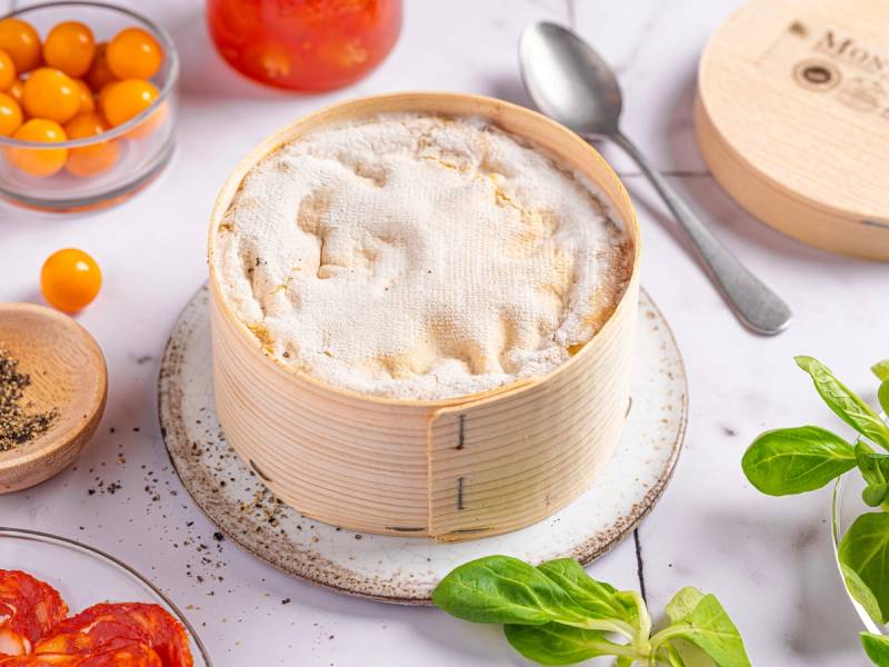 En rund bambuskål med fersk ost, omgitt av tomater, basilikablad og en skje, på et hvitt bord.